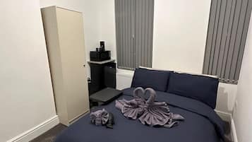 A hotel room with a bed, a wardrobe, a small table with a kettle, and a window with blinds.
