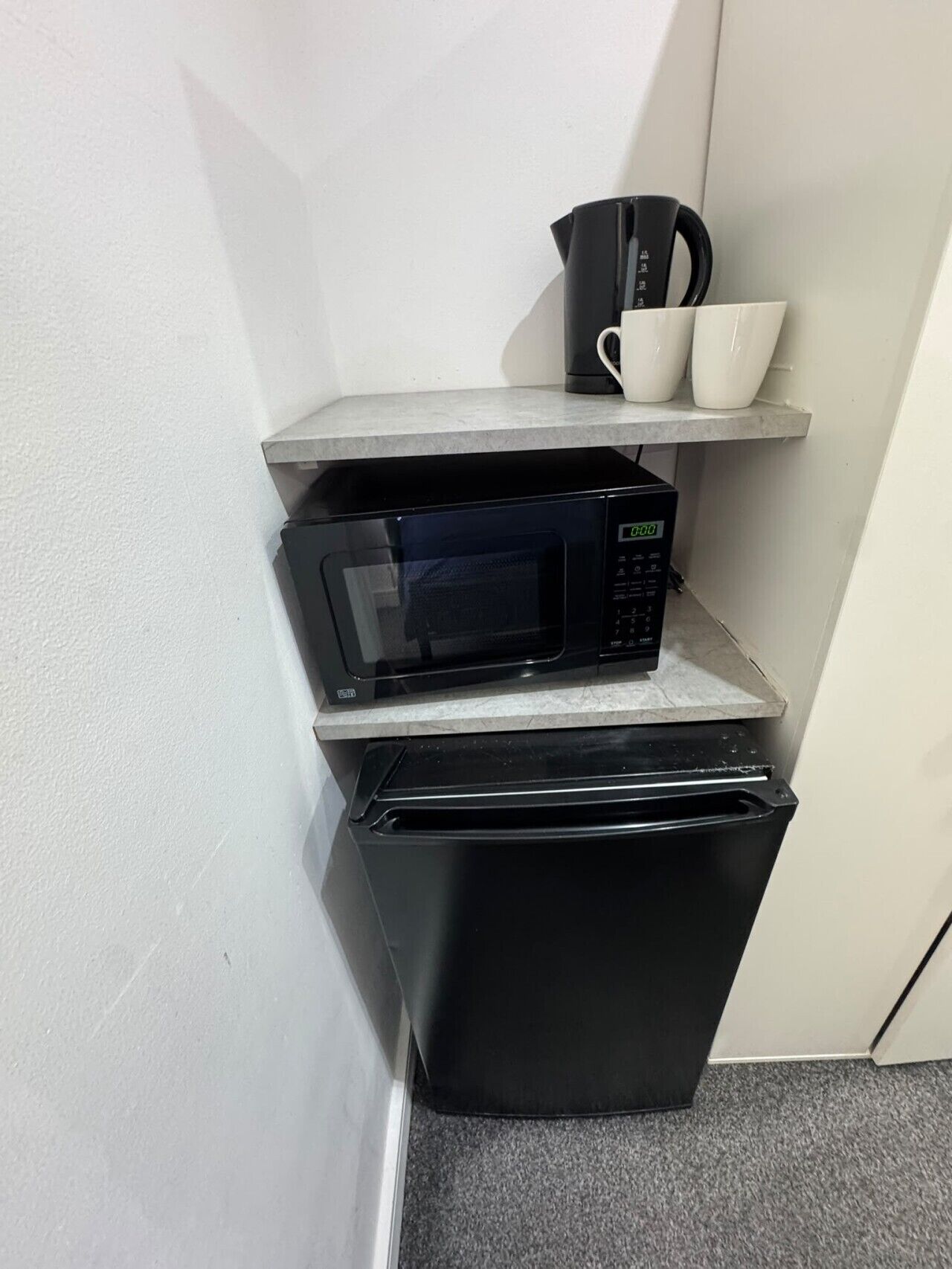 A compact kitchenette with a microwave, a small refrigerator, and a shelf with cups and a coffee maker.