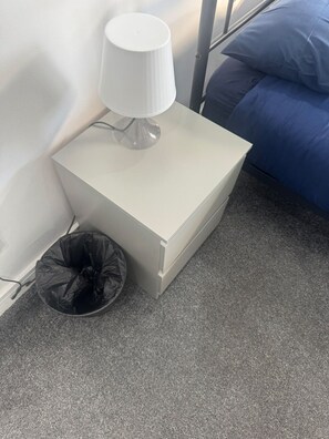 A white bedside table with a lamp, a black trash bin, and a blue bed in a room with a grey carpet. - London Victoria 15 minutes away (Greater London)