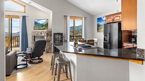 A modern kitchen with a stone fireplace, bar stools, and a view of mountains through large windows.