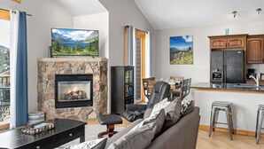 A living room with a stone fireplace, a television, a sofa, and a kitchen area with cabinets and a refrigerator.