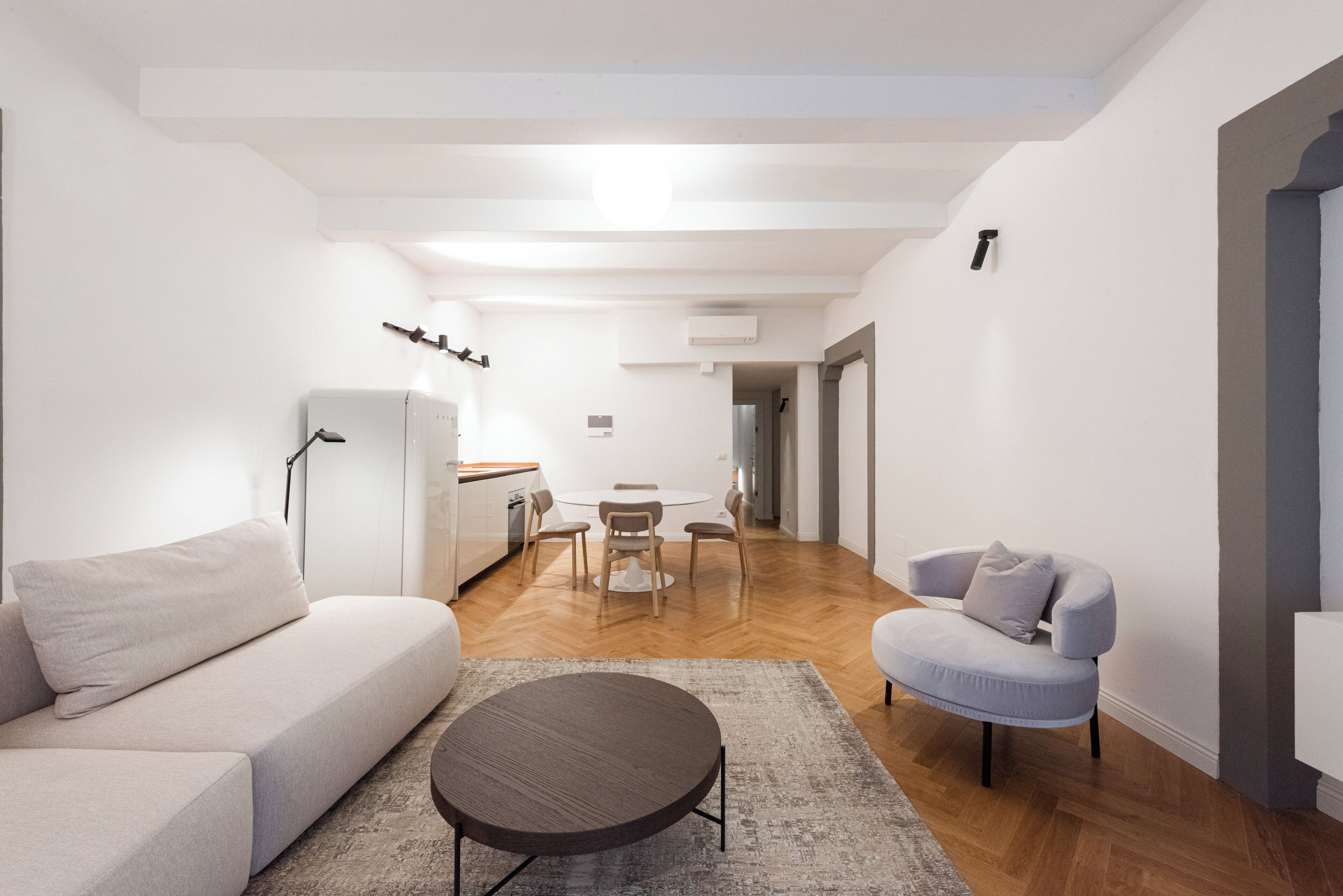 Un salon moderne avec un canapé, une table basse ronde et un fauteuil bleu clair. On aperçoit une table à manger avec des chaises à l’arrière-plan.