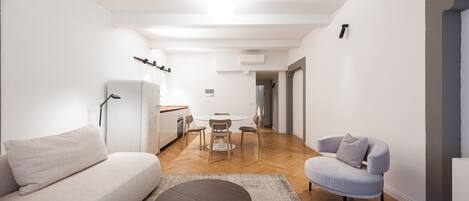 Un salon moderne avec un canapé, une table basse ronde et un fauteuil bleu clair. On aperçoit une table à manger avec des chaises à l’arrière-plan.