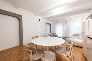 Ein modernes Wohnzimmer mit einem runden weißen Tisch, an dem vier Stühle stehen, sowie einer Couch und einem großen Spiegel an der Wand.