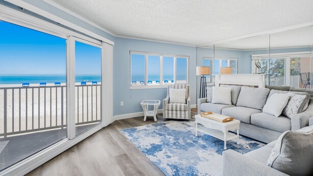 Ein geräumiges Wohnzimmer mit einer großen Schiebeglasdoor zum Balkon, ein graues Sofa, ein kleiner weißer Beistelltisch und ein blaues Teppich mit Muster.