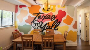 Un comedor con una mesa y sillas de madera, un mural en la pared con la inscripción 'Texas Y'all' y una araña.