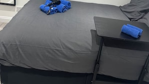 A single bed with a grey mattress and black headboard, a small black side table with a blue towel, and a poster on the wall.