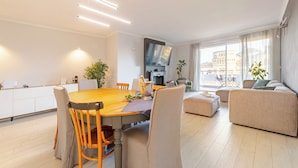 A modern living room with a wooden dining table, beige sofas, and a flat-screen TV mounted on the wall.