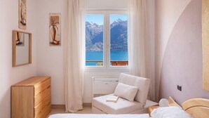 A bedroom with a bed, a wooden dresser, a window with curtains, and a view of mountains and water. - Mont&Lac | Lake View Apartment in Limone (Limone Sul Garda)