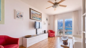 A spacious living room with a wooden floor, a ceiling fan, a red armchair, a white sofa, a TV, a fireplace, and a large window with a mountain view. - Mont&Lac | Lake View Apartment in Limone (Limone Sul Garda)