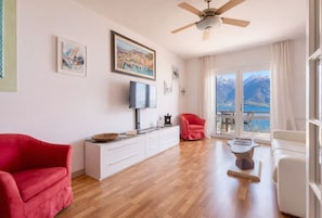 A spacious living room with a wooden floor, a ceiling fan, a red armchair, a white sofa, a TV, a fireplace, and a large window with a mountain view. - Mont&Lac | Lake View Apartment in Limone (Limone Sul Garda)