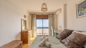 Ein Schlafzimmer mit einem Bett, einer Holzkommode, einer Bank, einem Fenster mit Vorhängen und einem Bild an der Wand.
