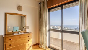 Ein Zimmer mit einem hölzernen Schminktisch, einem großen Spiegel, einem kleinen Tisch mit Pflanze und Blick auf die Stadt durch ein großes Fenster.