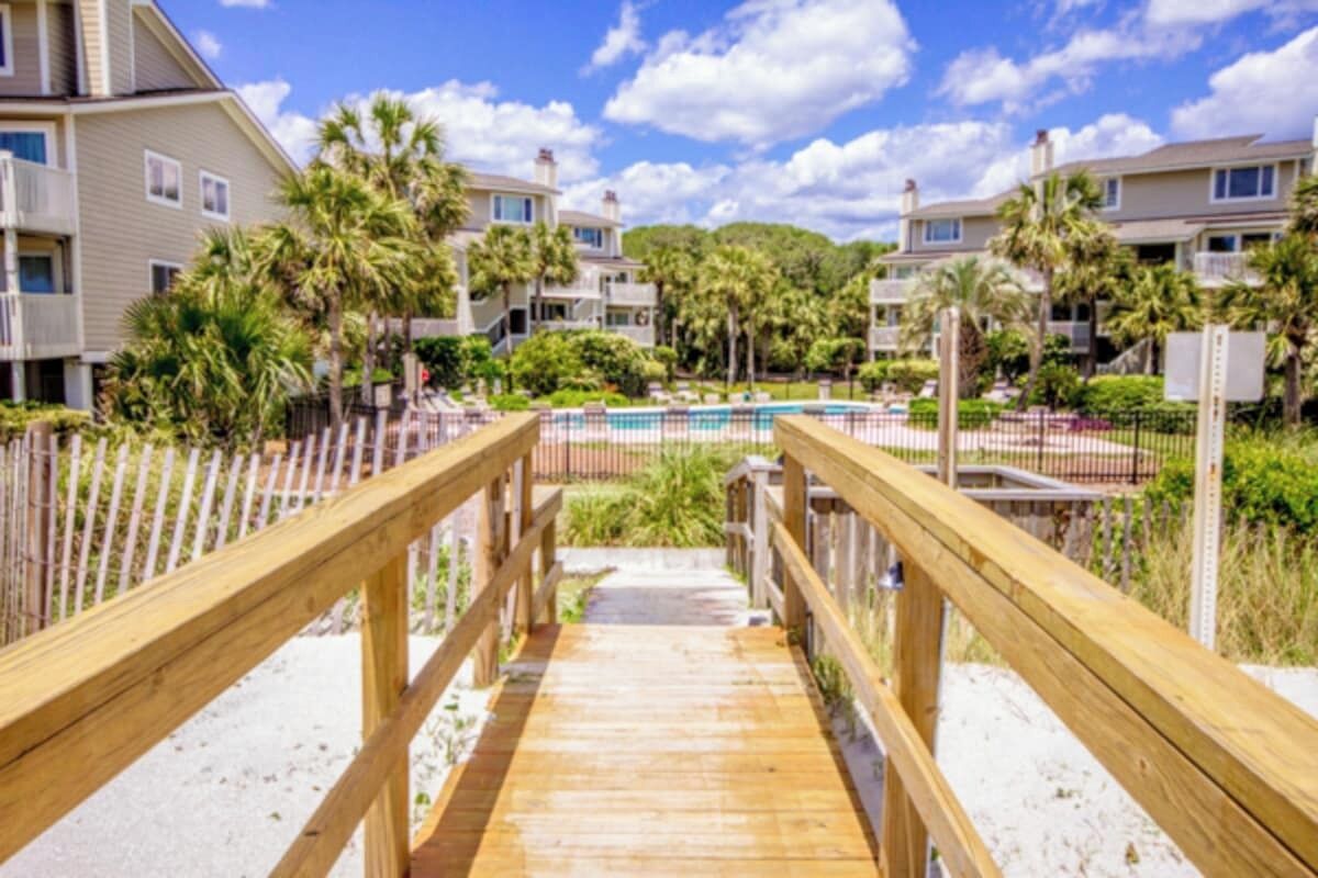 A wooden boardwalk leading to a pool area surrounded by palm trees and a fence.