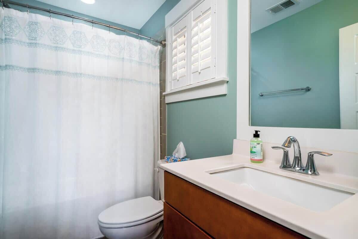 Ein Badezimmer mit Toilette, Duschvorhang, Waschbecken mit Spiegel und Fenster mit Jalousien.