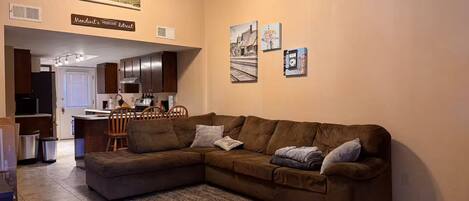 A living room with a brown sectional sofa, a small dining table, and a kitchen in the background.
