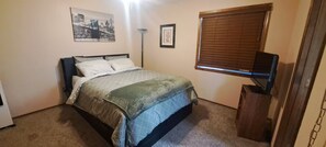 Een slaapkamer met een bed, een televisie op een dressoir, een raam met zonwering, en een foto aan de muur.