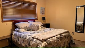 Een slaapkamer met een bed, een nachtkastje, een spiegel aan de muur en een raam met jaloezieën.