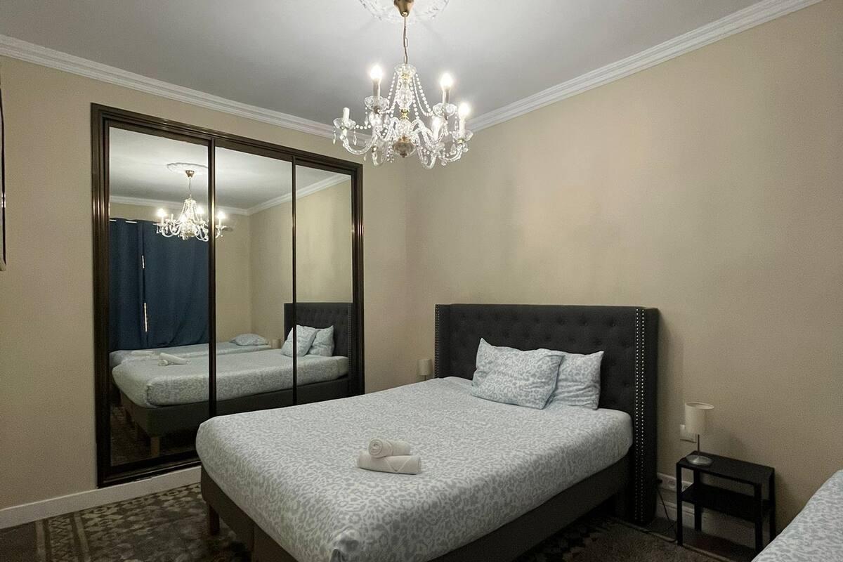 A hotel room with a large bed, a mirror, a chandelier, and a small bedside table.