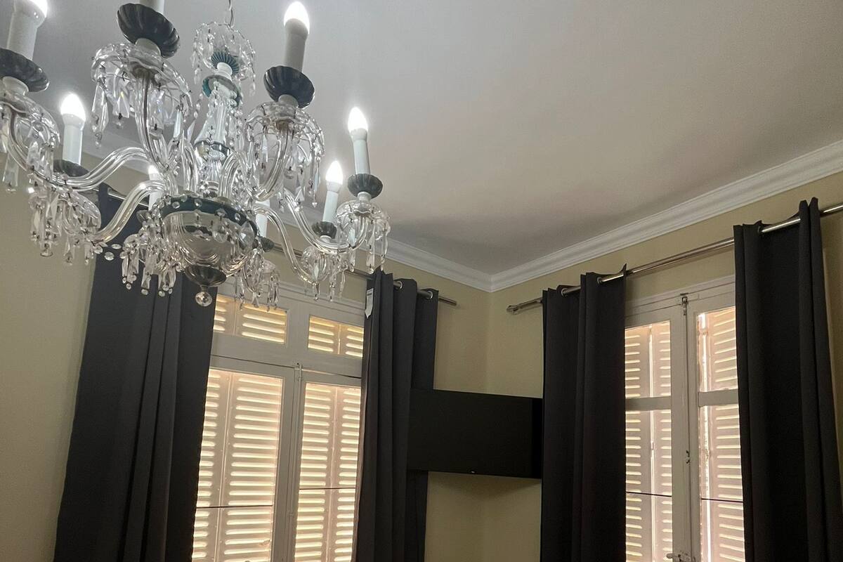 A bedroom with a chandelier, a bed with a patterned bedspread, and windows with blinds.