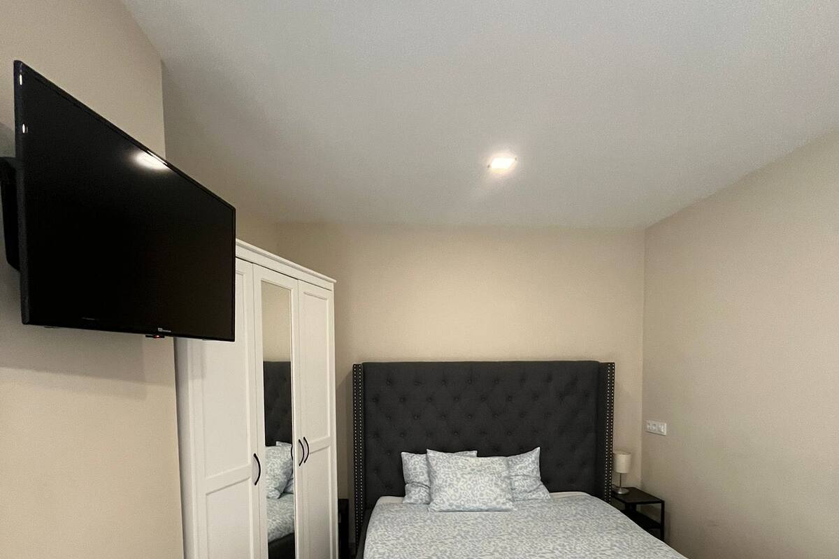 A bedroom with a bed, a TV mounted on the wall, a wardrobe, and a bedside table.