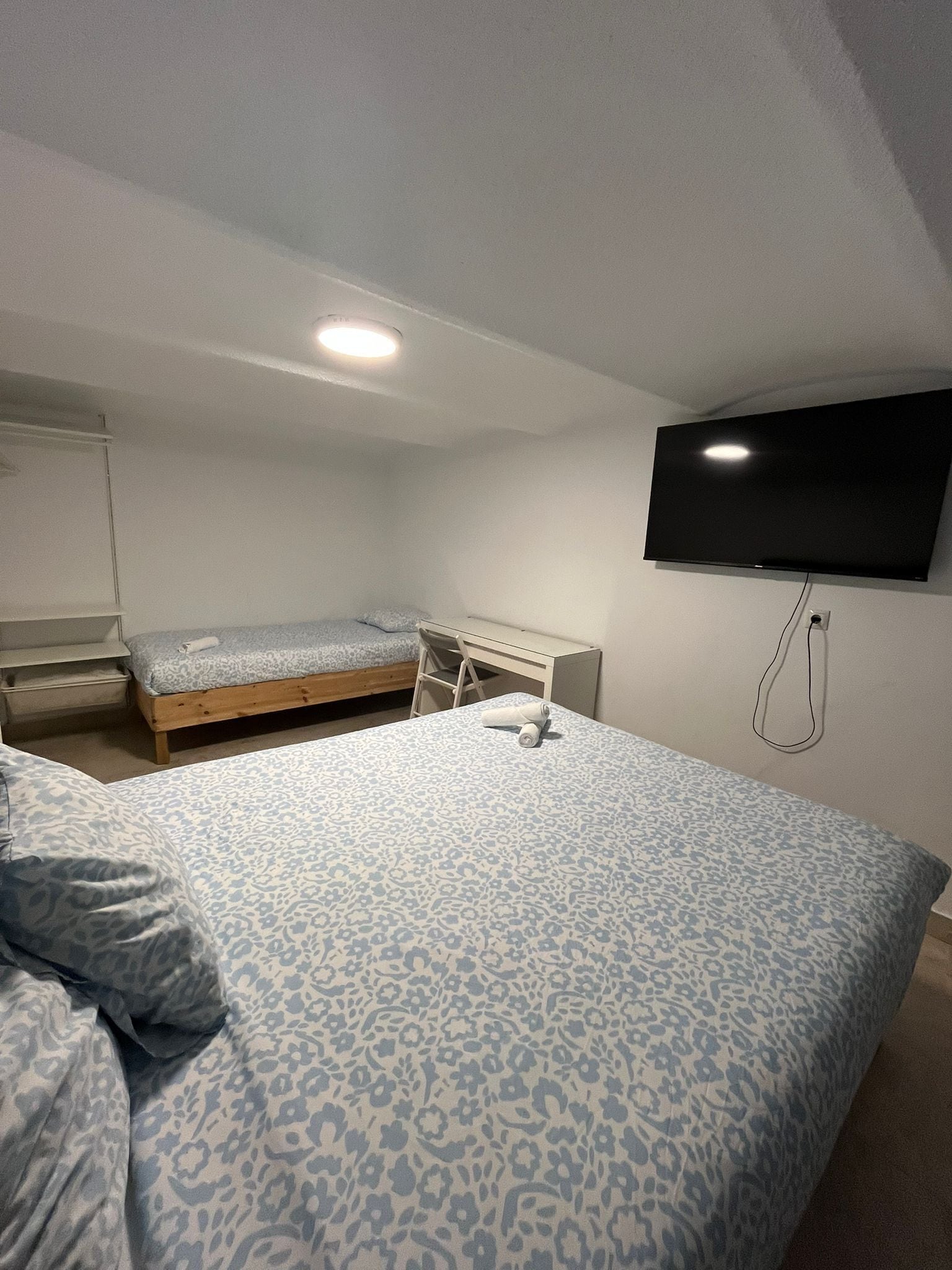 Un dormitorio con una cama grande, una cama más pequeña, un escritorio, una silla, un televisor montado en la pared y una lámpara en el techo.