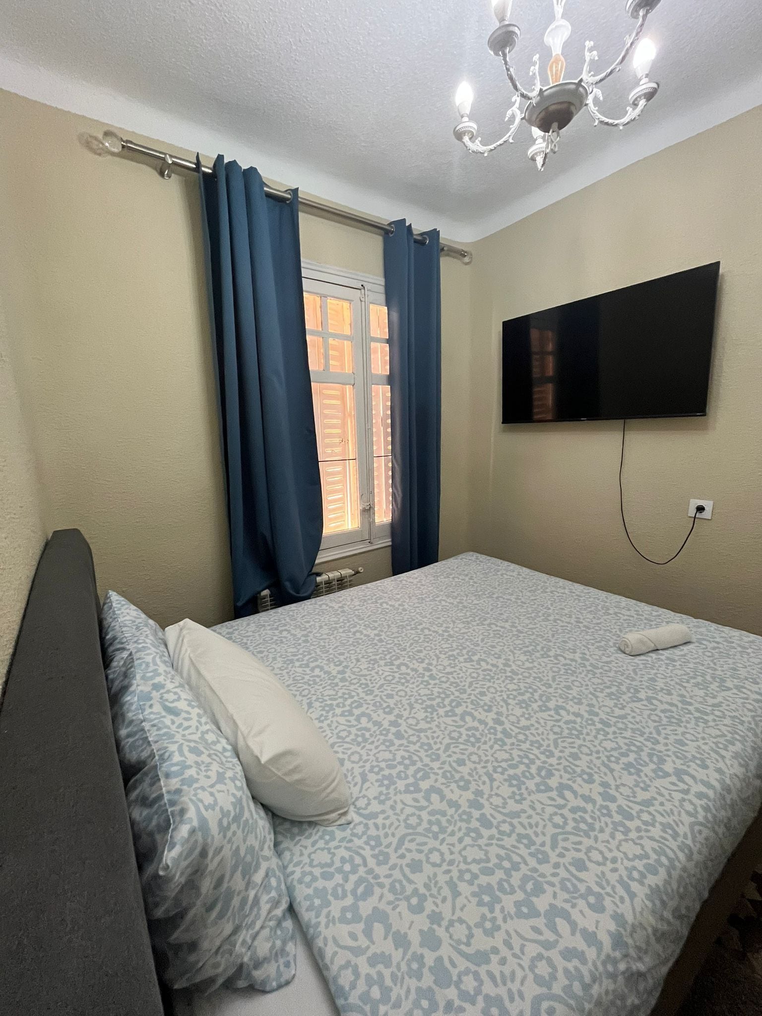 Une chambre à coucher comprenant un lit, un téléviseur à écran plat fixé au mur, une fenêtre avec des rideaux et un lustre.