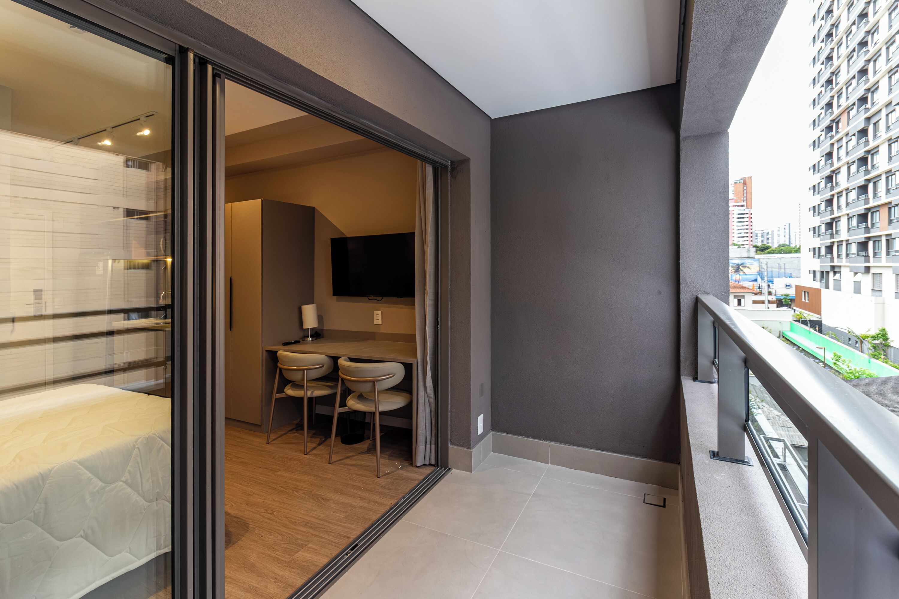 Un appartement moderne doté d’un balcon, d’un coin repas avec deux chaises, d’un téléviseur à écran plat fixé au mur et offrant une vue sur le paysage urbain.