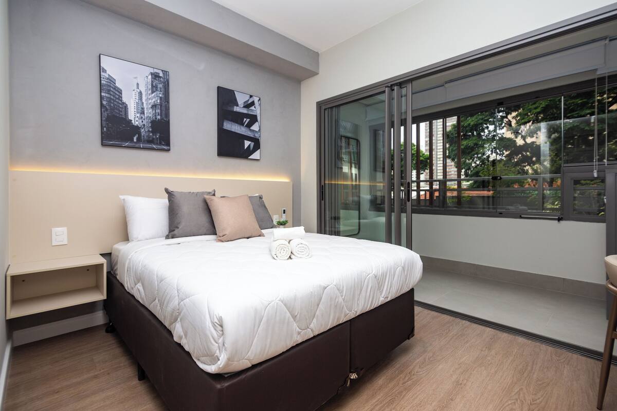 Un dormitorio moderno con una cama grande, una mesita de noche, un estante en la pared y un ventanal corredero que da a un balcón.