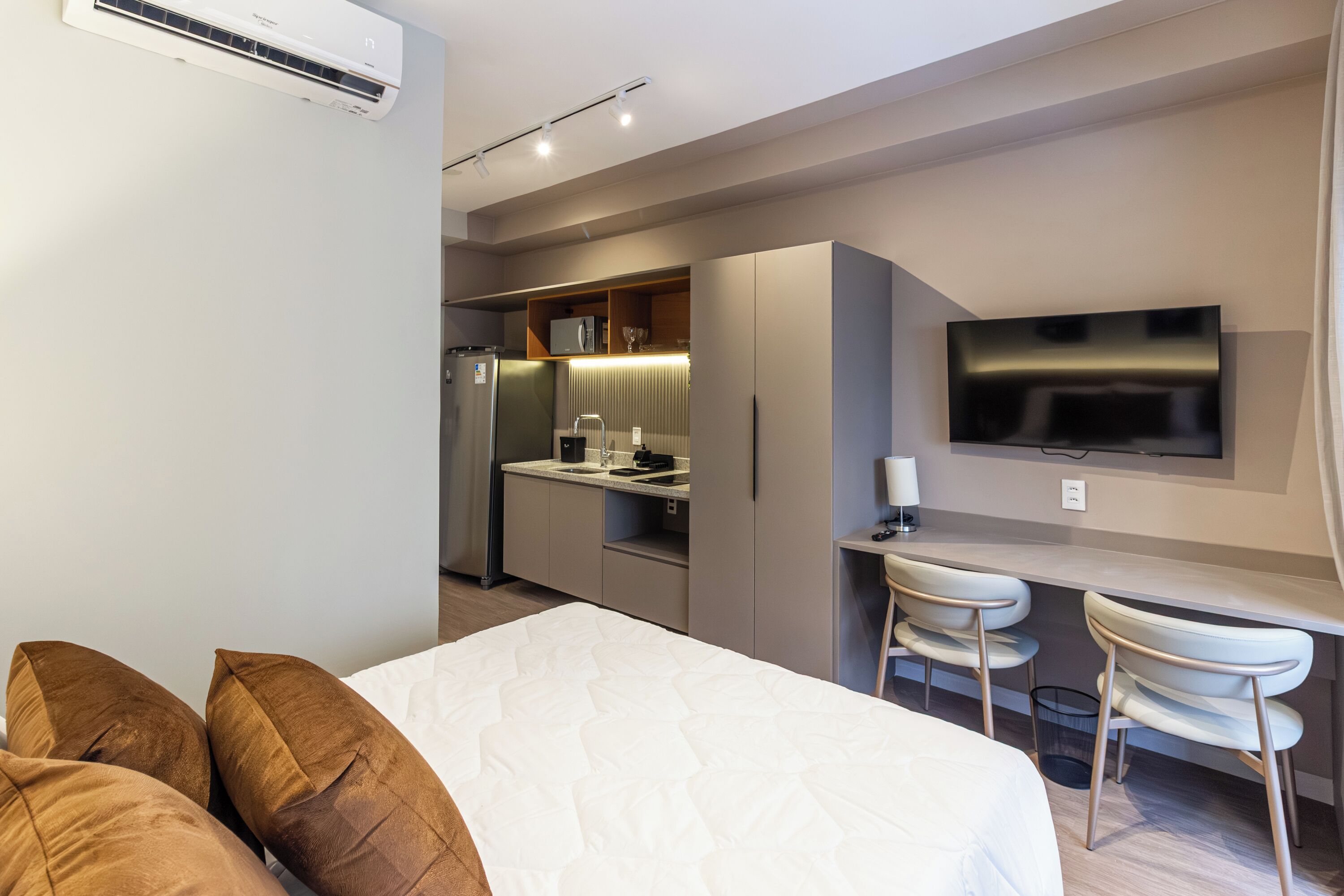 A modern hotel room with a bed, a desk, a TV, and a kitchenette.
