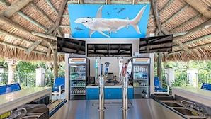 A beachside bar with a thatched roof, a large shark mural, and a counter with a cash register and bar tools. - Accessible 2BR Pool-View Family Suite in Naples (Naples)