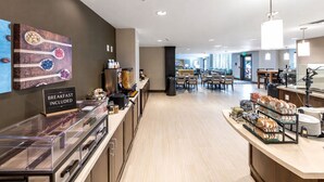 Un área de desayuno moderna en un hotel, con un letrero que indica desayuno incluido, una mesa de buffet, una zona de estar y vistas del comedor.