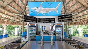 Un bar en la playa con techo de paja, un gran mural de un tiburón y una barra con caja registradora y utensilios de bar.