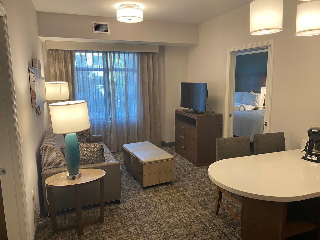 A hotel room with a sofa, a small table with a lamp, a TV on a stand, a dining table, and a bed with a headboard.