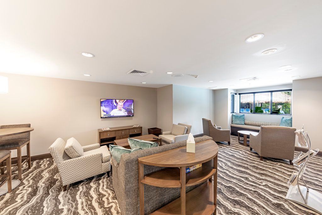 A modern hotel room with a large flat-screen TV, comfortable seating, a patterned carpet, and a window with a view.