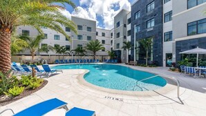 Piscina con sillas reclinables azules, palmeras y un edificio moderno de varios pisos y grandes ventanales.