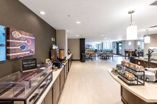 A modern hotel breakfast area with a sign indicating breakfast is included, a buffet counter, seating area, and a view of the dining space.