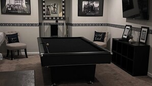 Ein Billardtisch in einem Raum mit einer Couch, einem an der Wand befestigten Fernseher und gerahmten Bildern.