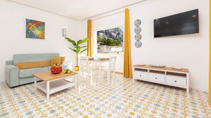 Una sala de estar con sofá, mesa de centro, mesa de comedor y sillas, televisor montado en la pared, y un balcón con vistas.