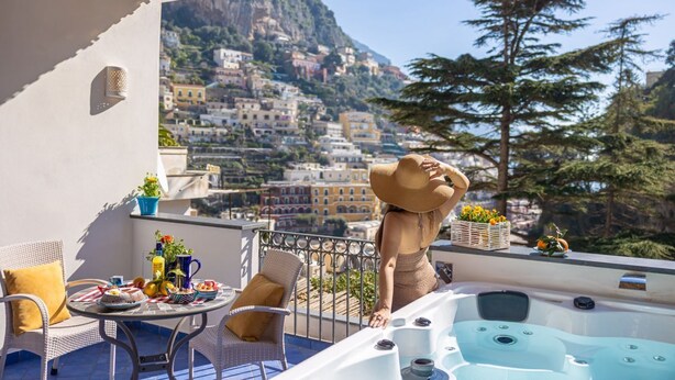 Un balcón con vistas a un pueblo en la ladera, muebles de mimbre, un jacuzzi y una mesa con comida y bebidas.