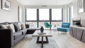 Uma sala de estar moderna com um sofá cinzento, uma poltrona azul, uma mesa de centro branca e uma TV sobre um armário de madeira.