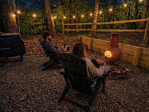 Dos personas sentadas al aire libre por la noche, con un horno de leña y luces colgantes de fondo.