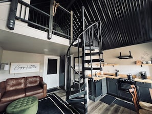 A loft-style living space with a staircase, a kitchenette, a brown leather sofa, and a green ottoman.