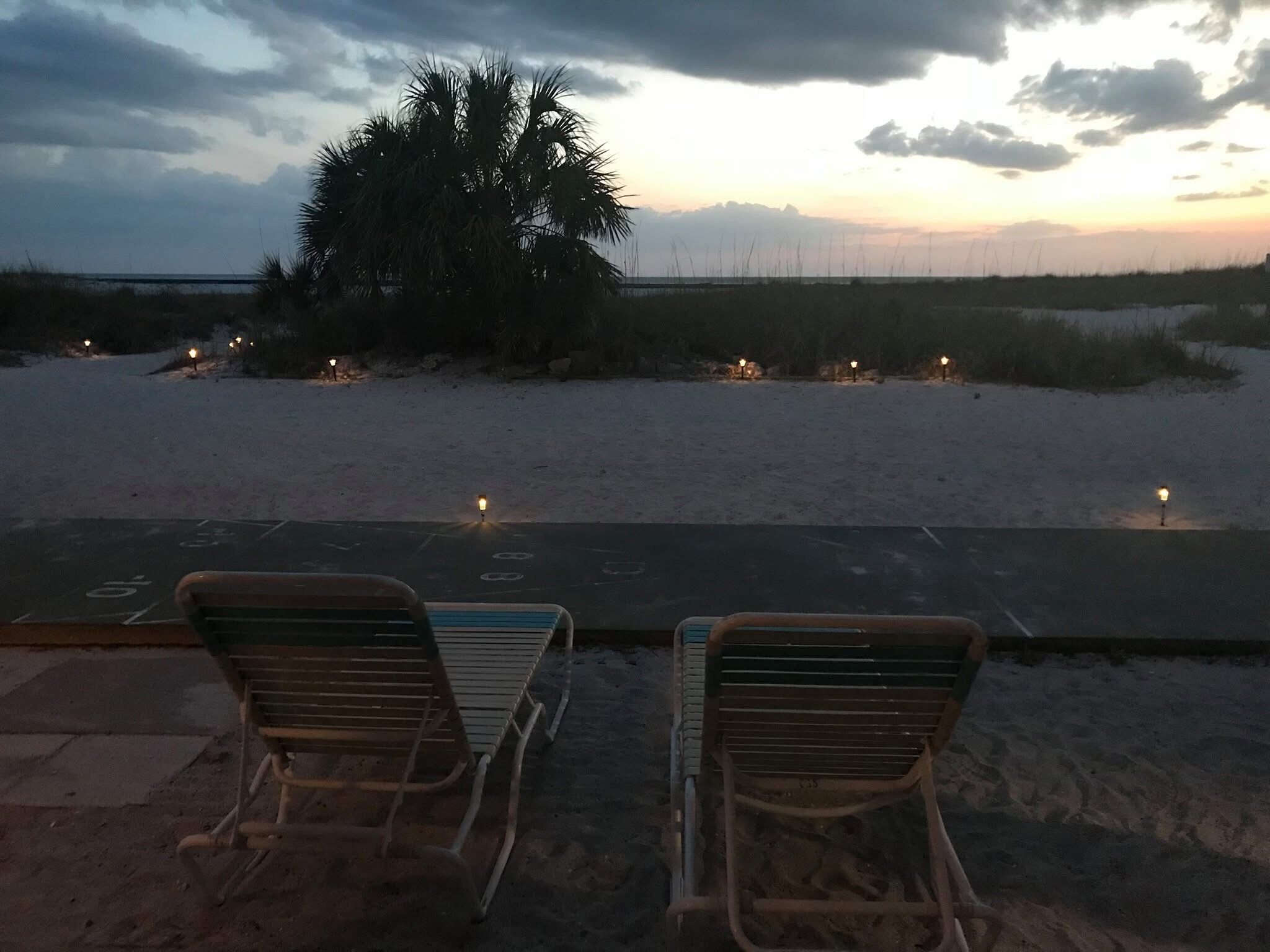 Dos sillas de descanso mirando una playa con una palmera y la costa a lo lejos, bajo un cielo crepuscular.