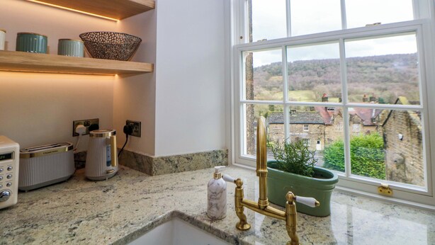 Eine Küche mit einer Granitarbeitsplatte, einem Messingarmaturen, einer grünen Pflanze und Blick auf ein Dorf durch ein großes Fenster.