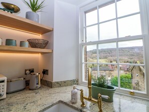 A kitchen with a granite countertop, a brass faucet, a green plant, and a view of a village through a large window. - The Reading Room (Hope Valley)