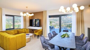 Una sala de estar moderna con un sofá amarillo, una mesa de comedor con sillas, un televisor de pantalla plana y grandes ventanales con vistas a la ciudad.