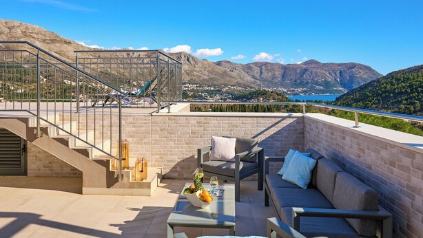 Una terraza en la azotea con barandilla de cristal, un sofá, una mesa con frutero y vistas a montañas y un lago.