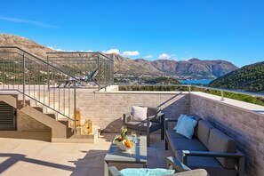 A rooftop terrace with a glass balustrade, a sofa, a table with a fruit bowl, and a view of mountains and a lake. - Hedera Estate, Villa Hedera IX (Zupa dubrovacka)