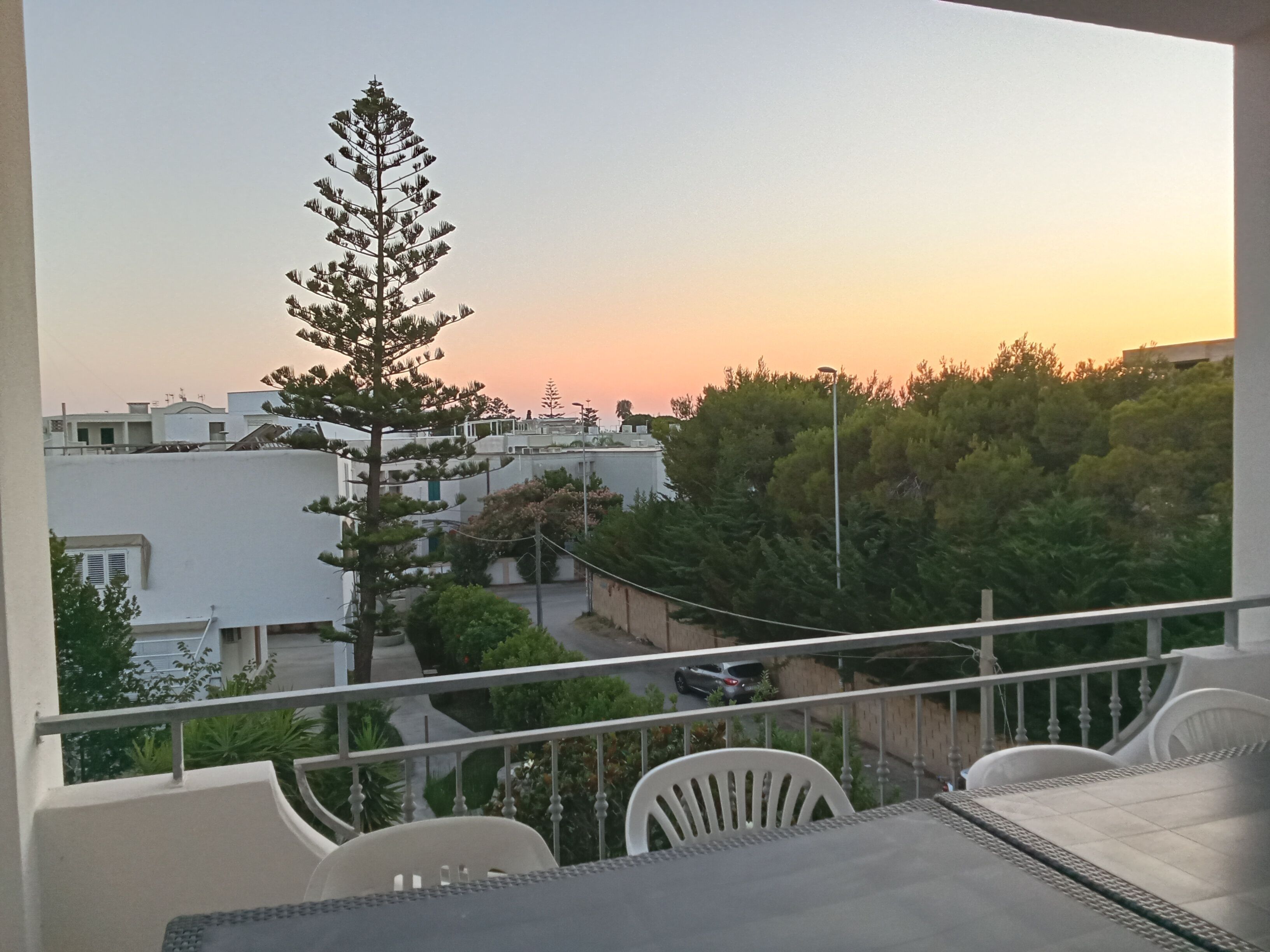 Un balcon avec une table et des chaises, donnant sur un quartier résidentiel avec des maisons et des arbres, au moment du coucher du soleil.